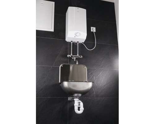 Wandmontierter AEG Warmwasserspeicher mit AEG Logo, verchromter Armatur, Edelstahl-Waschbecken und Siphon an einer Fliesenwand.
