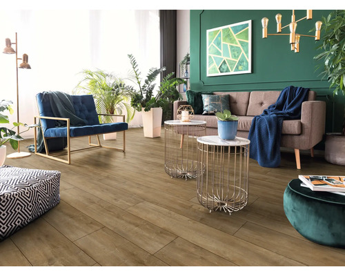 Salon moderne avec sol aspect bois, canapé marron, fauteuil bleu, tables d'appoint dorées et mur vert avec plantes d'intérieur.