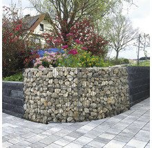 Gabion courbé rempli de pierres naturelles utilisé comme mur de jardin, entouré de plantes fleuries et d'un chemin pavé.