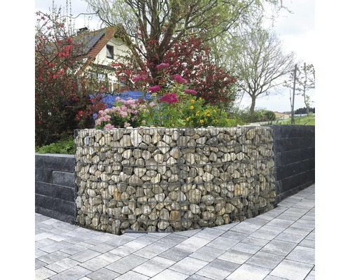 Gabion courbé rempli de pierres naturelles utilisé comme mur de jardin, entouré de plantes fleuries et d'un chemin pavé.
