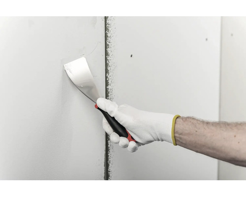 Une main gantée utilise un couteau à enduire pour appliquer de l'enduit sur une plaque de plâtre.