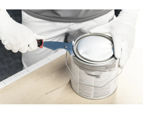Personne ouvrant un pot de peinture avec un grattoir à peinture multifonction en métal sur un support en bois.