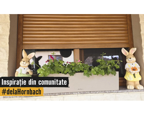 Balkonkasten mit Geranien und zwei Hasenfiguren auf einer Fensterbank vor einem Holz-Rollladen.