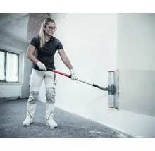 Femme utilisant une spatule à lisser large avec perche télescopique pour lisser un mur dans une pièce lumineuse.