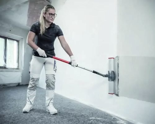 Femme utilisant une spatule à lisser large avec perche télescopique pour lisser un mur dans une pièce lumineuse.