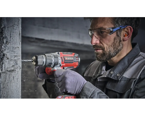 Artisan utilisant une perceuse visseuse à percussion sans fil Einhell pour percer un mur en béton, portant lunettes et protection auditive.