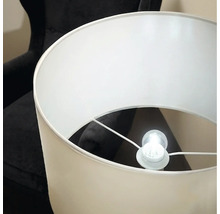 Un lampadaire élégant avec un abat-jour cylindrique et une ampoule allumée devant un fauteuil sombre.