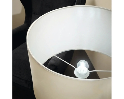 Un lampadaire élégant avec un abat-jour cylindrique et une ampoule allumée devant un fauteuil sombre.