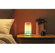 Lampe de table LED cylindrique avec dégradé de couleurs sur une table de chevet à côté d'un lit avec des livres et une plante.