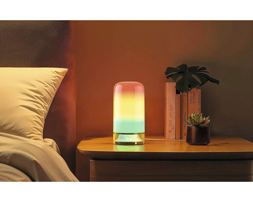Lampe de table LED cylindrique avec dégradé de couleurs sur une table de chevet à côté d'un lit avec des livres et une plante.