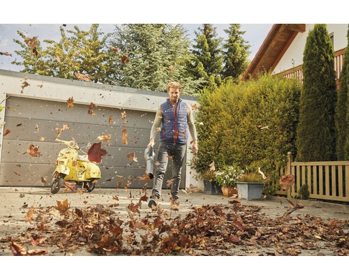 Ein Mann benutzt einen Laubsauger, um Laub auf einer Auffahrt zu entfernen. Im Hintergrund befinden sich eine Garage und ein Motorroller.
