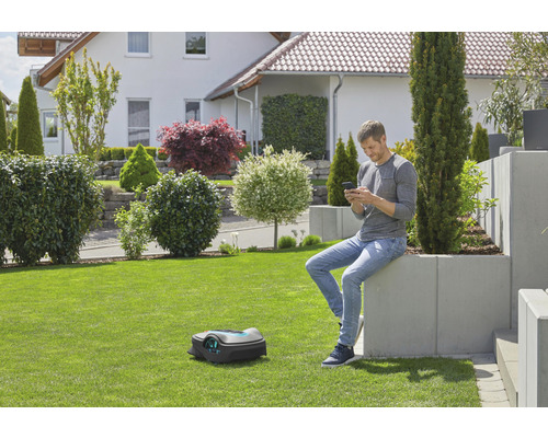 Ein Mann sitzt im Garten und bedient einen Rasenmähroboter mit seinem Mobiltelefon.