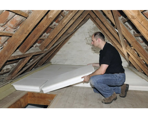 Mann verlegt Dämmplatten auf einem Dachboden mit Holzbalken.