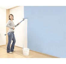 Une femme peint un mur avec un rouleau à peinture monté sur une perche télescopique, un seau de peinture est posé sur le sol.