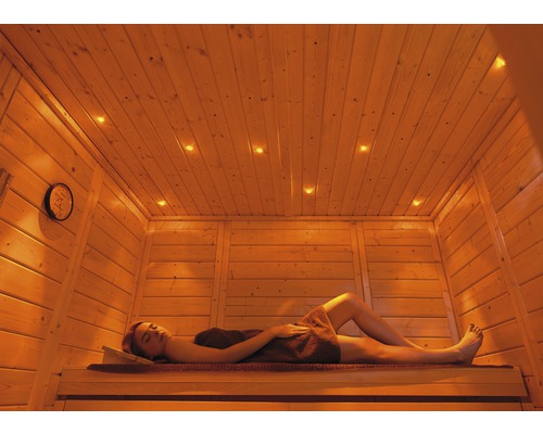 Kit de visions colorées pour sauna A Weka, avec module de commande Femme se détend dans un sauna en bois avec horloge et éclairage au plafond.
