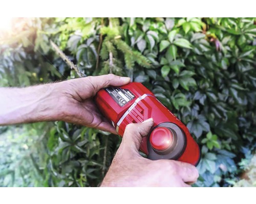 Person bedient eine Akku-Grasschere vor einer grünen Hecke.