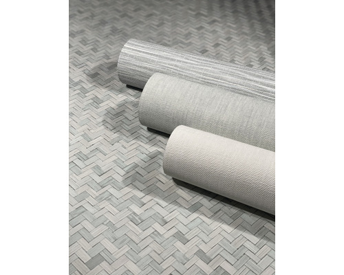 Drei Tapetenrollen mit unterschiedlichen Mustern liegen auf einem geflochtenen Untergrund.