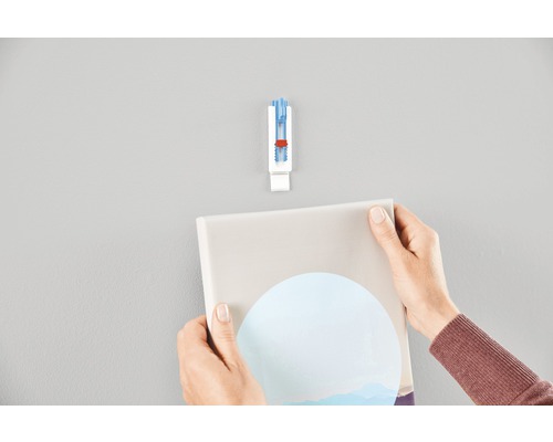 Eine Person hängt mit einem selbstklebenden Bilderhaken ein Bild an die Wand.