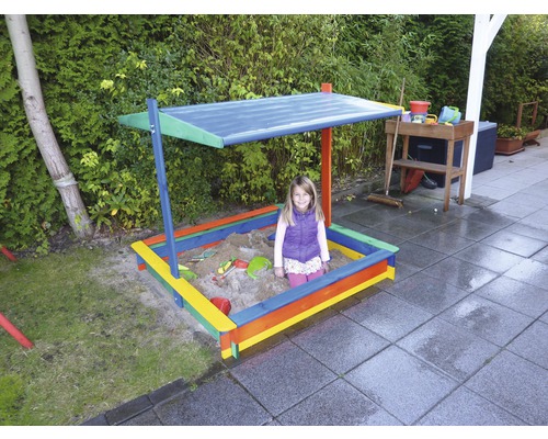 Un enfant est assis dans un bac à sable coloré avec un toit pare-soleil dans le jardin.