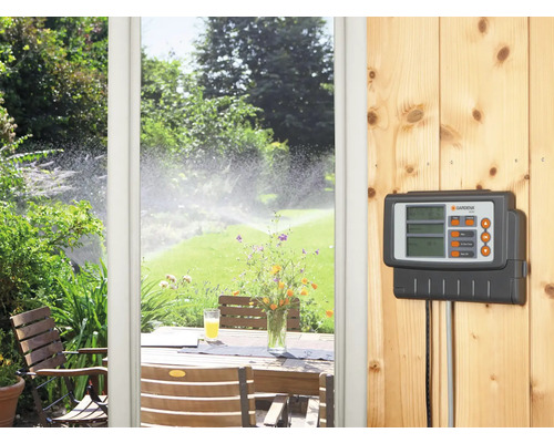 Gardena Bewässerungssteuerung an einer Holzwand, im Hintergrund eine Terrasse und ein Rasensprenger