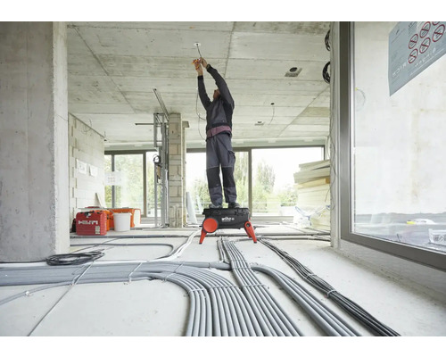 Elektriker steht auf einem Tritt und arbeitet in einem Rohbau an der Decke. Auf dem Boden liegen viele Kabel.