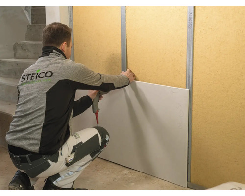 Ein Mann befestigt eine weiße Gipskartonplatte an einer Wand mit Metallprofilen und Holzwerkstoffplatten.